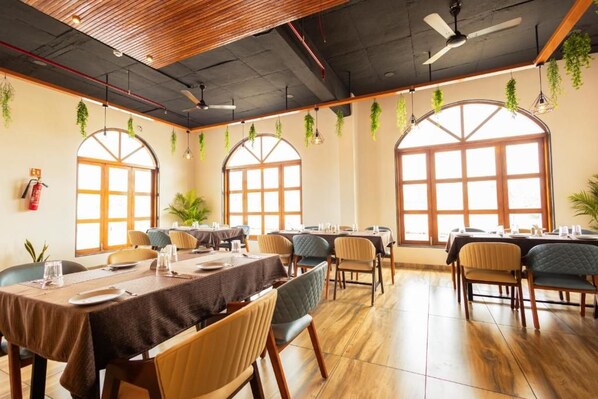 A dining area with tables set for service, large windows with arched tops, and hanging plants.