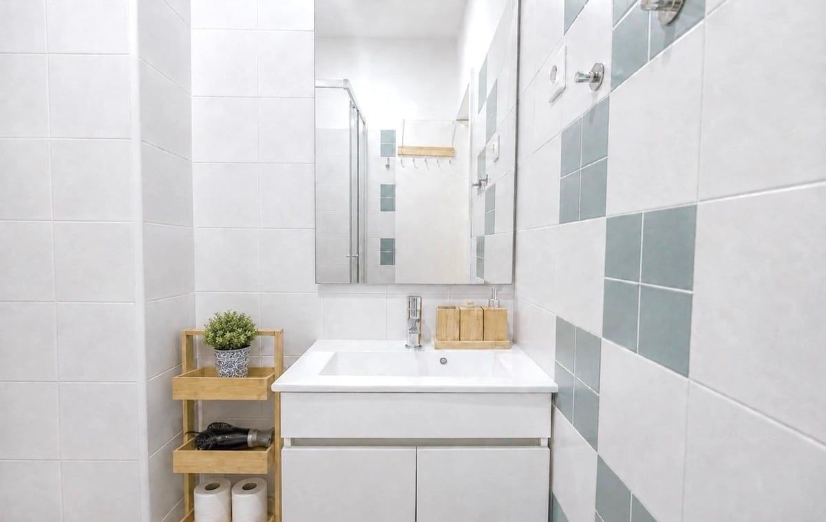 Une petite salle de bain aux murs recouverts de carreaux blancs, avec un lavabo blanc, un miroir et une étagère en bois où sont rangés les articles de toilette.