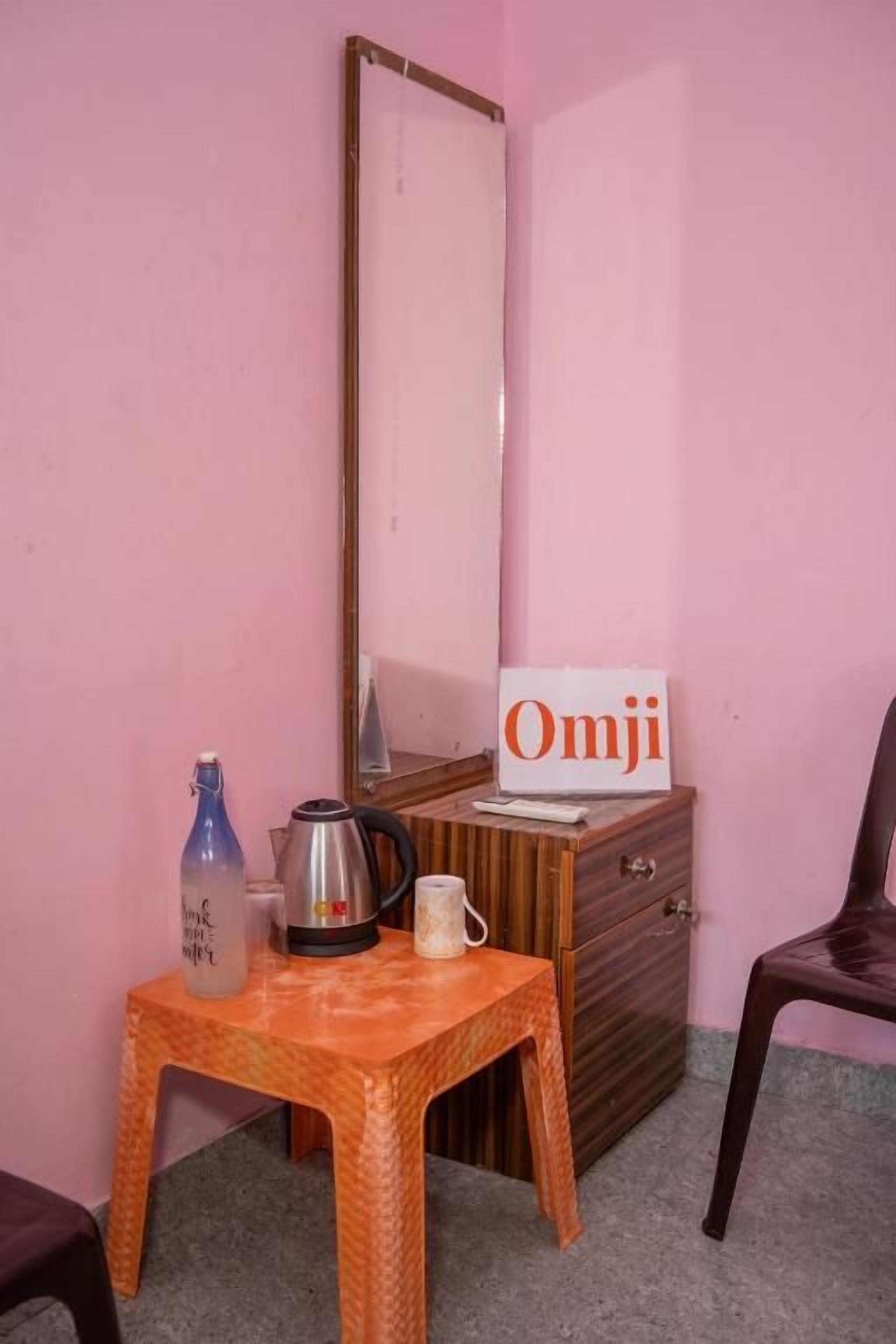 A room with a pink wall, a wooden dresser, a mirror, a small table with a kettle, a mug, and a bottle.