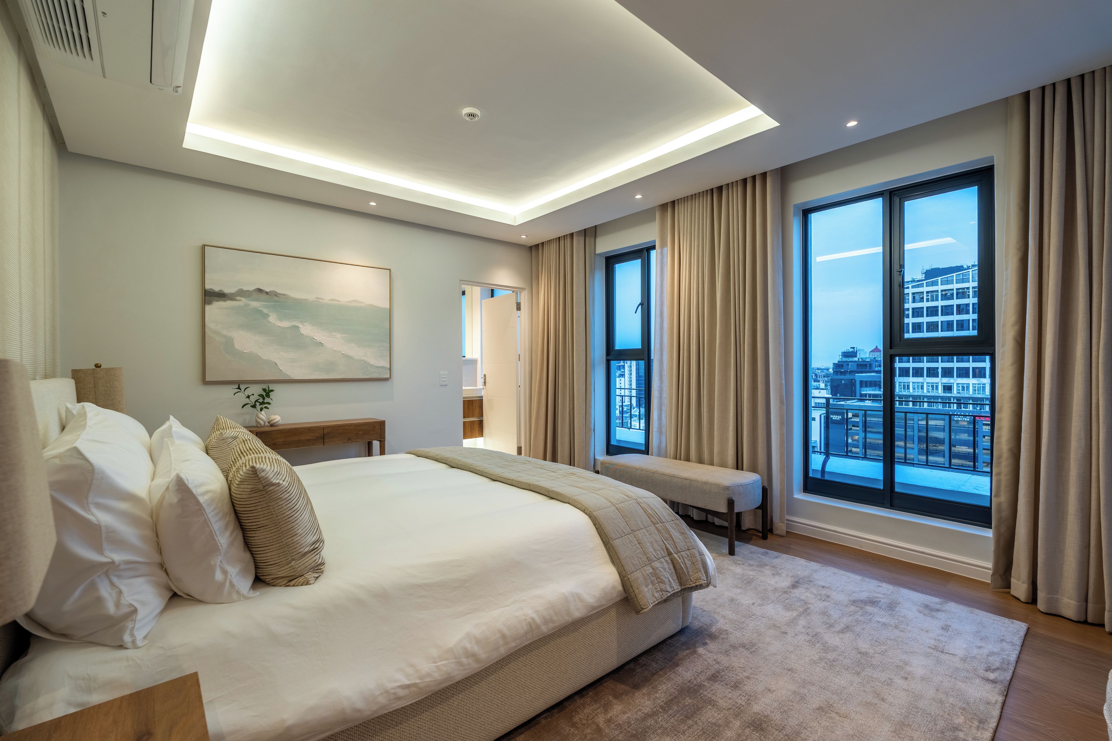 A modern hotel room with a large bed, a bedside table, a bench, a large window with curtains, and a painting on the wall.