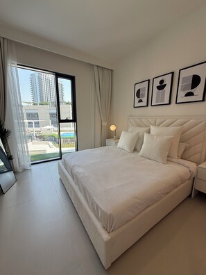 A hotel room with a large bed, a nightstand, a lamp, and a view of a swimming pool and buildings. - 1-BHK - Dubai Creek Harbor - Pool View (Dubai)