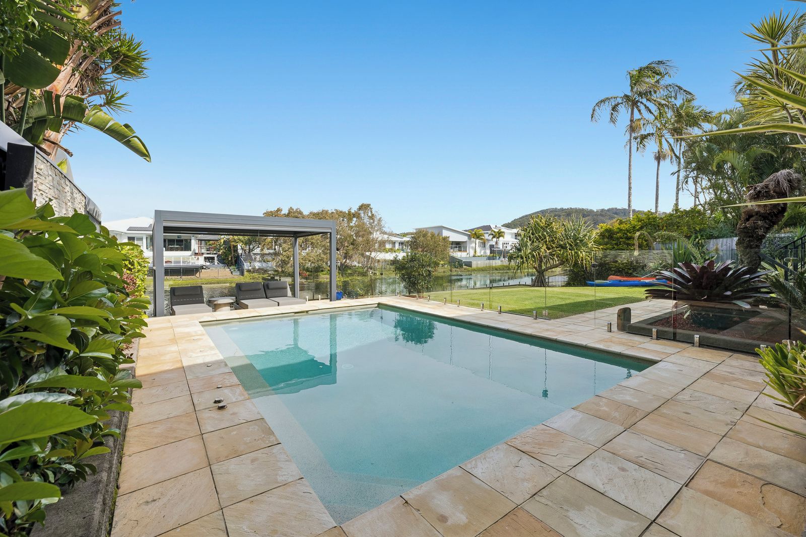 A rectangular swimming pool with a covered seating area, surrounded by a landscaped garden with palm trees and a view of a residential area.