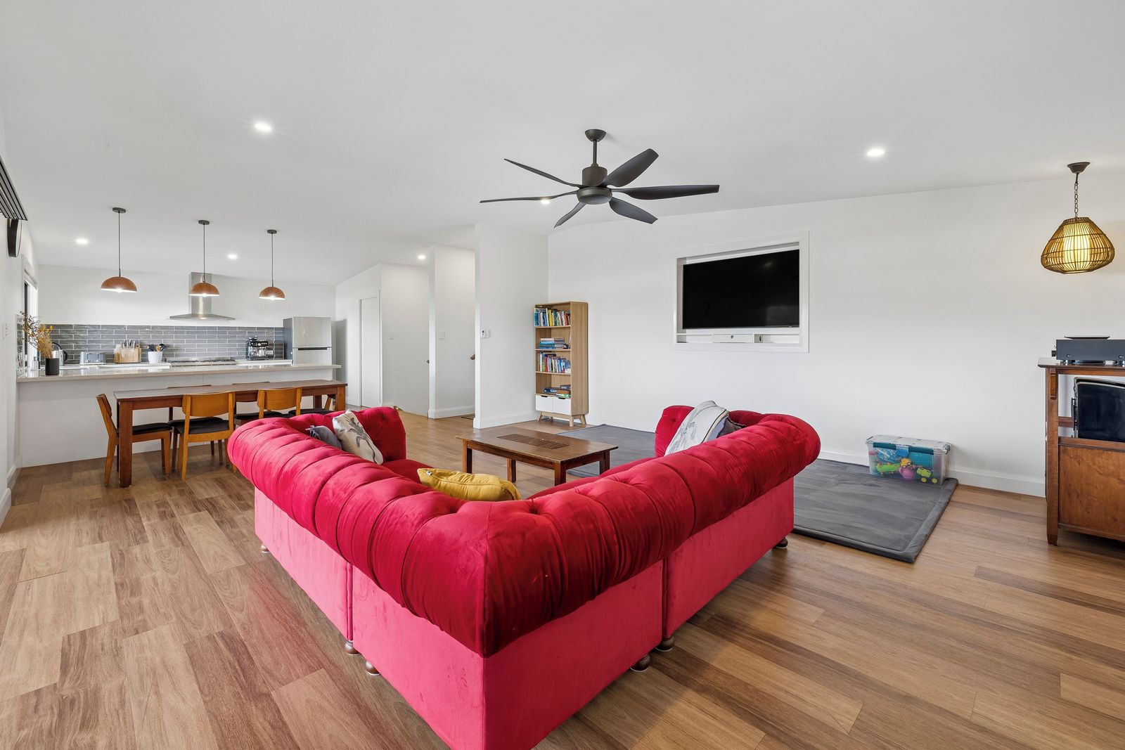 A modern living room with a red sofa, wooden flooring, a ceiling fan, pendant lights, a flat-screen TV, and a kitchen area in the background.