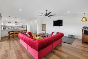 Ein modernes Wohnzimmer mit einem roten Sofa, Holzboden, Deckenventilator, Pendelleuchten, einem Flachbildfernseher und einer Küchenzeile im Hintergrund.