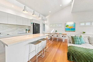 A modern kitchen with a central island, bar stools, a dining area with a table and chairs, and a living room with a sofa and decorative pillows. - Paradise Palms | Walk to Beach | Boat Parking (Agnes Water)