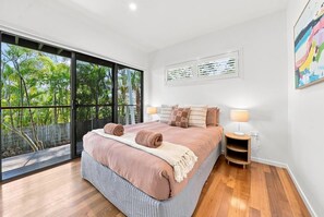 A bedroom with a large bed, a nightstand, a window with a view of trees, and a painting on the wall. - Paradise Palms | Walk to Beach | Boat Parking (Agnes Water)