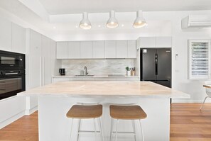 A modern kitchen with white cabinetry, a marble countertop, and a central island with bar stools. - Paradise Palms | Walk to Beach | Boat Parking (Agnes Water)