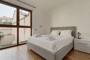A bedroom with a large window, a bed with white linens, a bedside table with a lamp, and a view of buildings outside. - GuestHost - Bright Apartment with a Balcony near the MM3 (Milan)