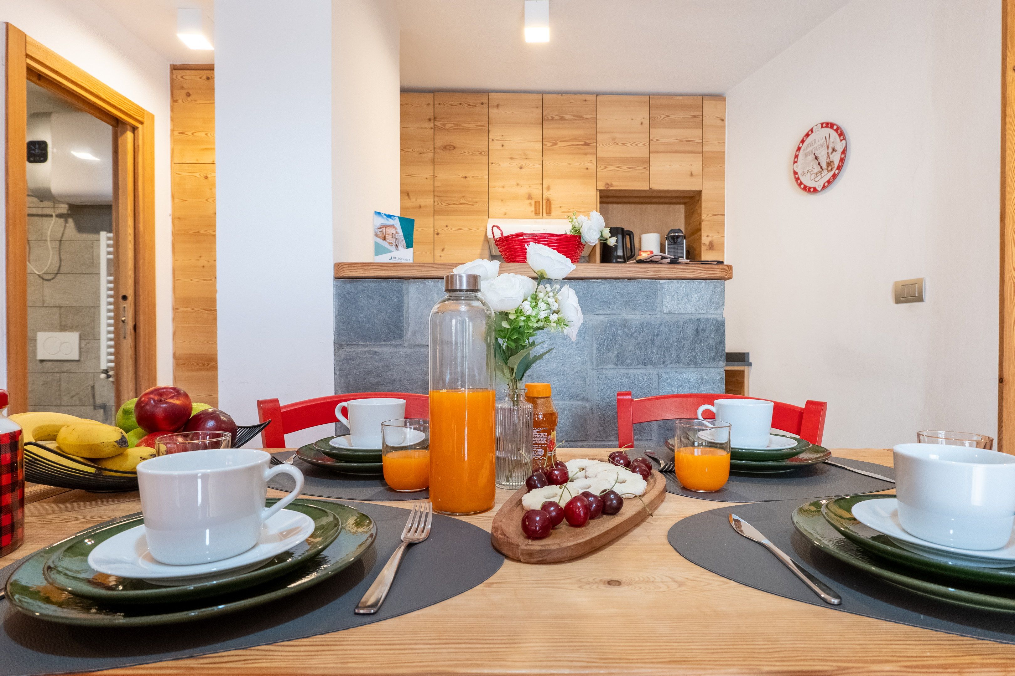 A breakfast setting with a wooden table, chairs, and a variety of fruits and beverages.