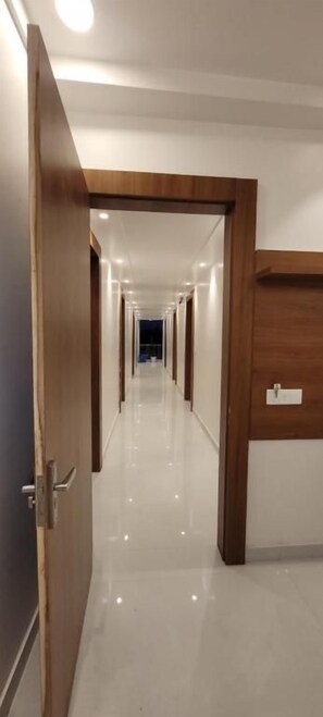 A hotel corridor with a wooden door on the left, a wooden wall on the right, and a polished floor. - Bhakt Bhagwat Kunj (Vrindavan)