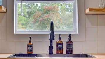 A kitchen sink with a black faucet, three bottles of dish soap, and a window with blinds.