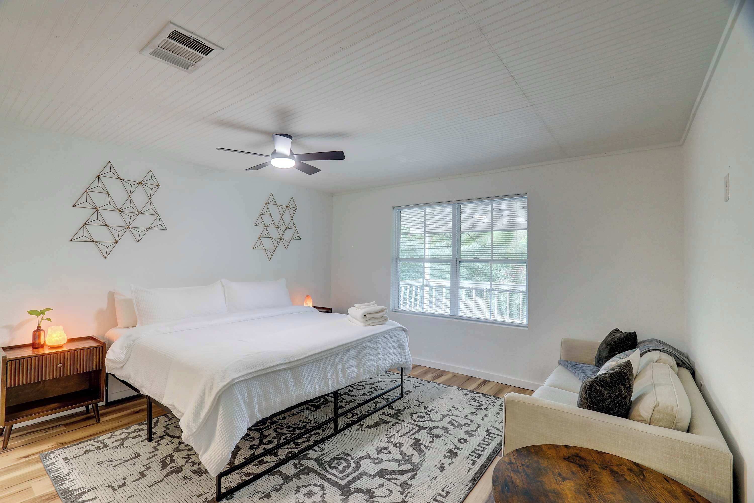 Une chambre à coucher comprenant un lit, une table de chevet, une lampe, une décoration murale aux motifs géométriques, un ventilateur de plafond, une fenêtre et une chaise.