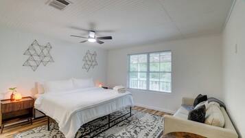 A bedroom with a bed, bedside table, lamp, geometric wall art, ceiling fan, window, and a chair.
