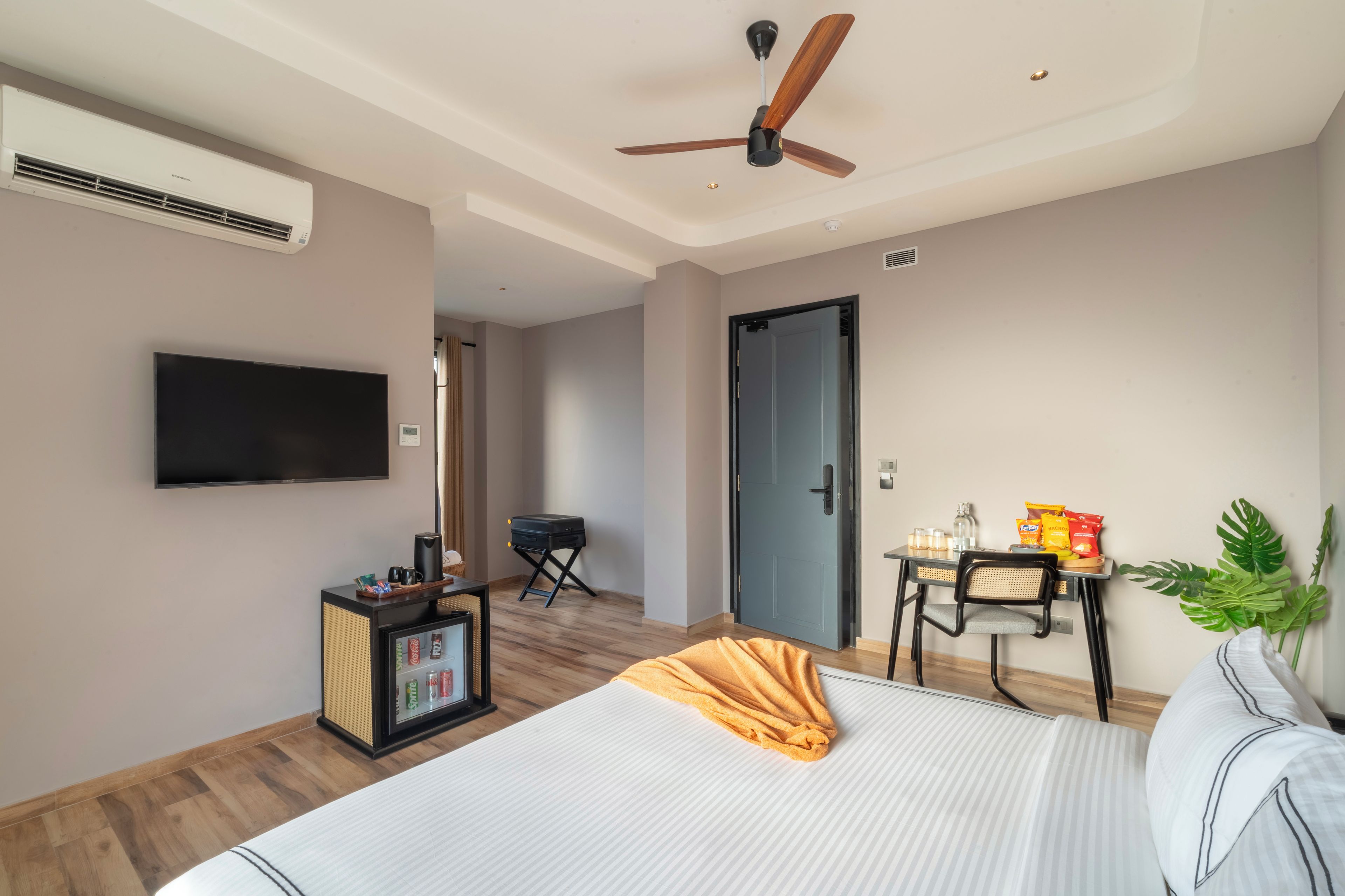Habitación de hotel con cama, televisor, ventilador de techo, una mesita con refrigerios y una planta en maceta.