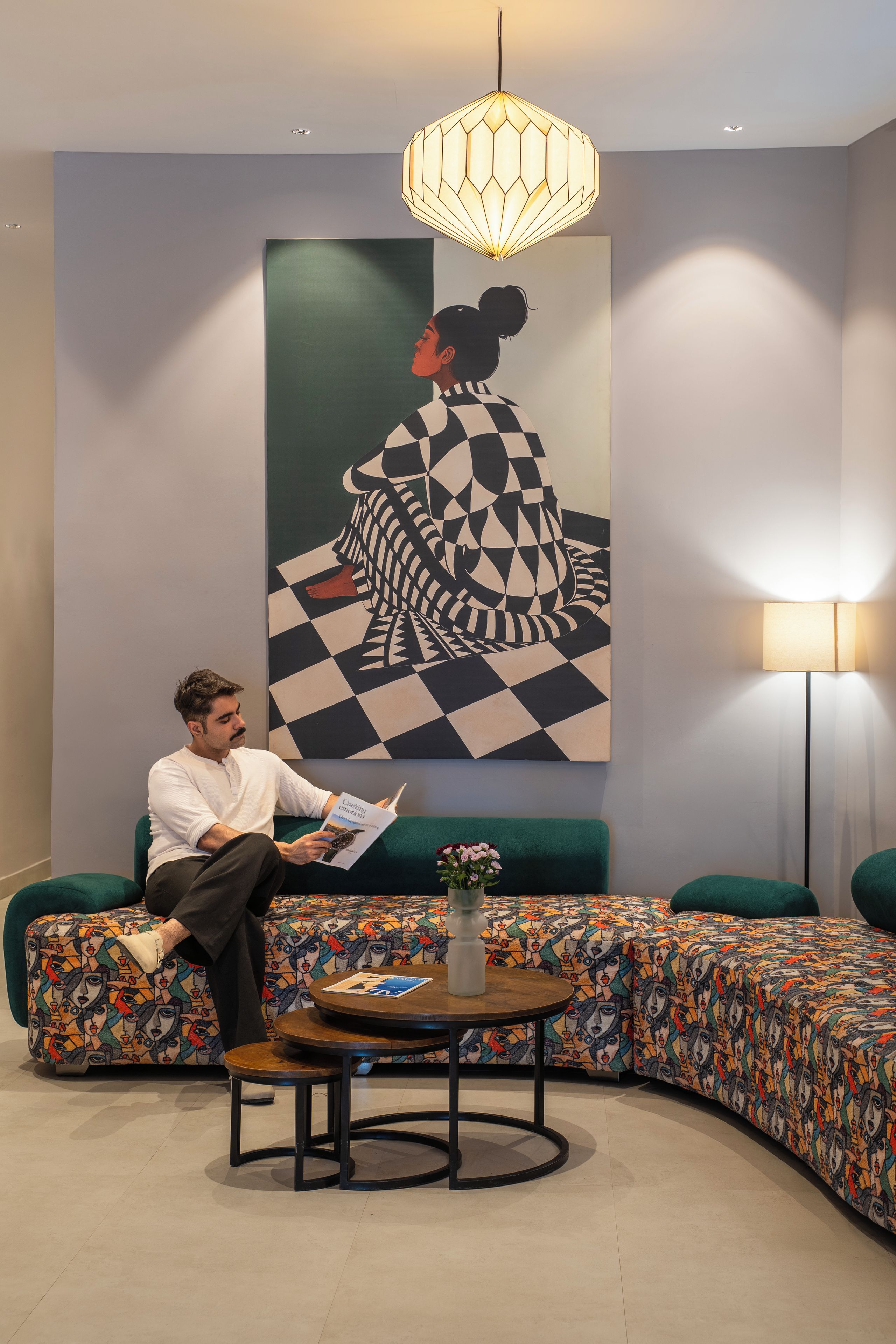 A person sitting on a green sofa reading a book in a room with a large abstract painting, a round table, and a floor lamp.