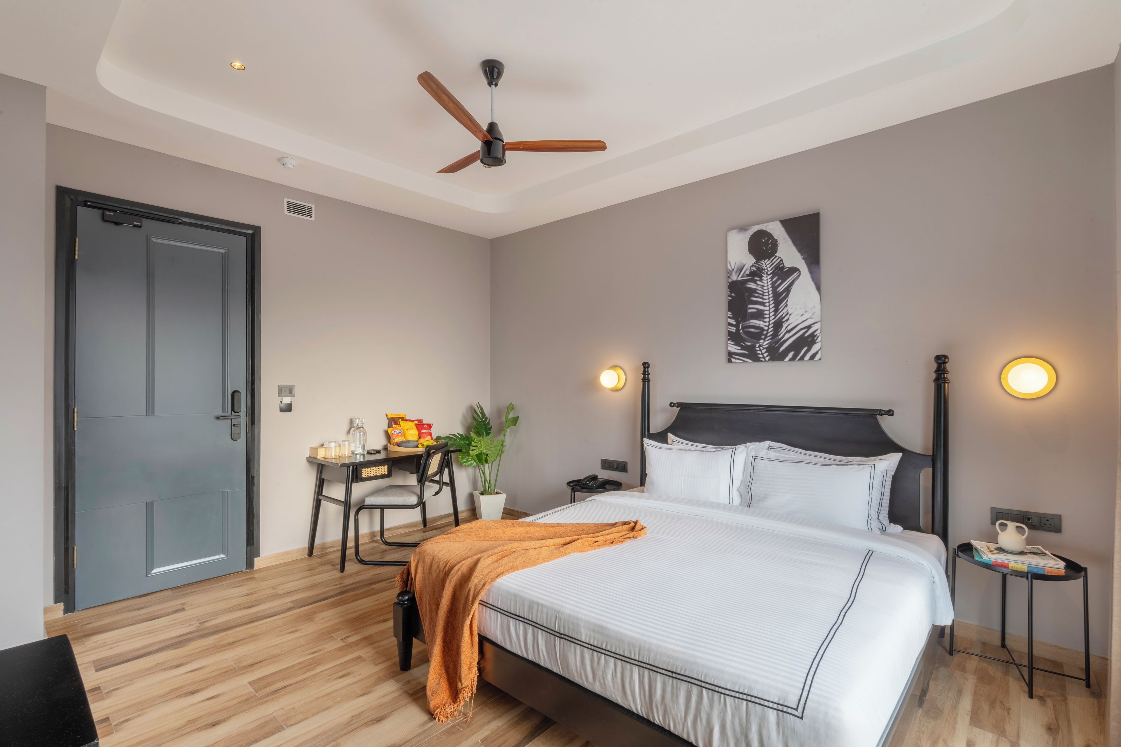 Una habitación de hotel con cama, mesitas de noche, un escritorio con silla, un ventilador de techo y una puerta.