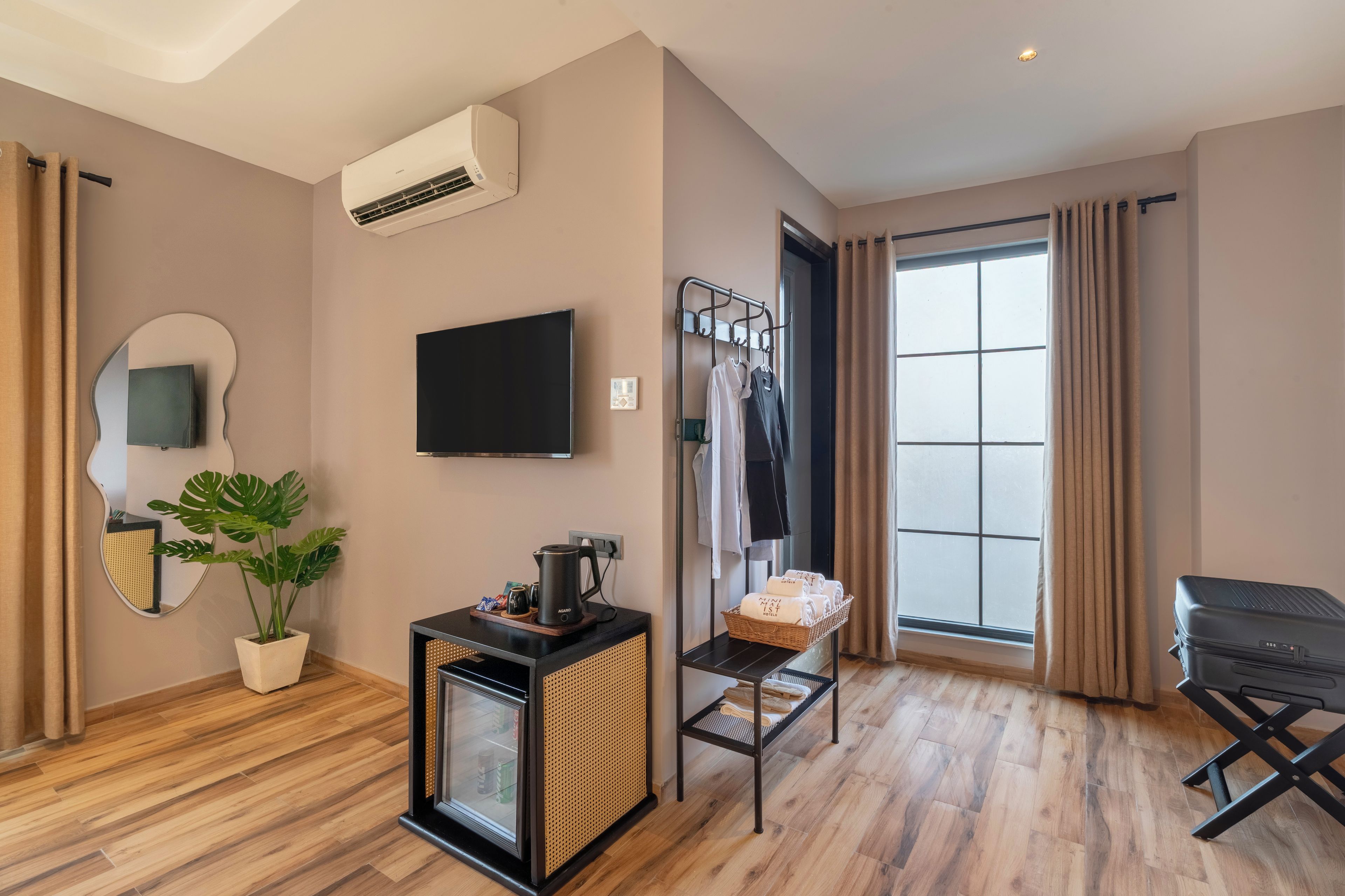 Habitación de hotel moderna con televisor de pantalla plana, planta en maceta, cafetera, minibar, zona de estar con mesa y sillas, y un ventanal con cortinas.