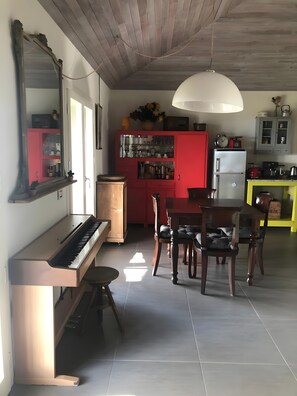 A room with a piano, dining table, chairs, a red cabinet, and a kitchen area with a refrigerator and cabinets. - Villa 'Cubetto' with Wi-Fi and Air Conditioning (Cesena)
