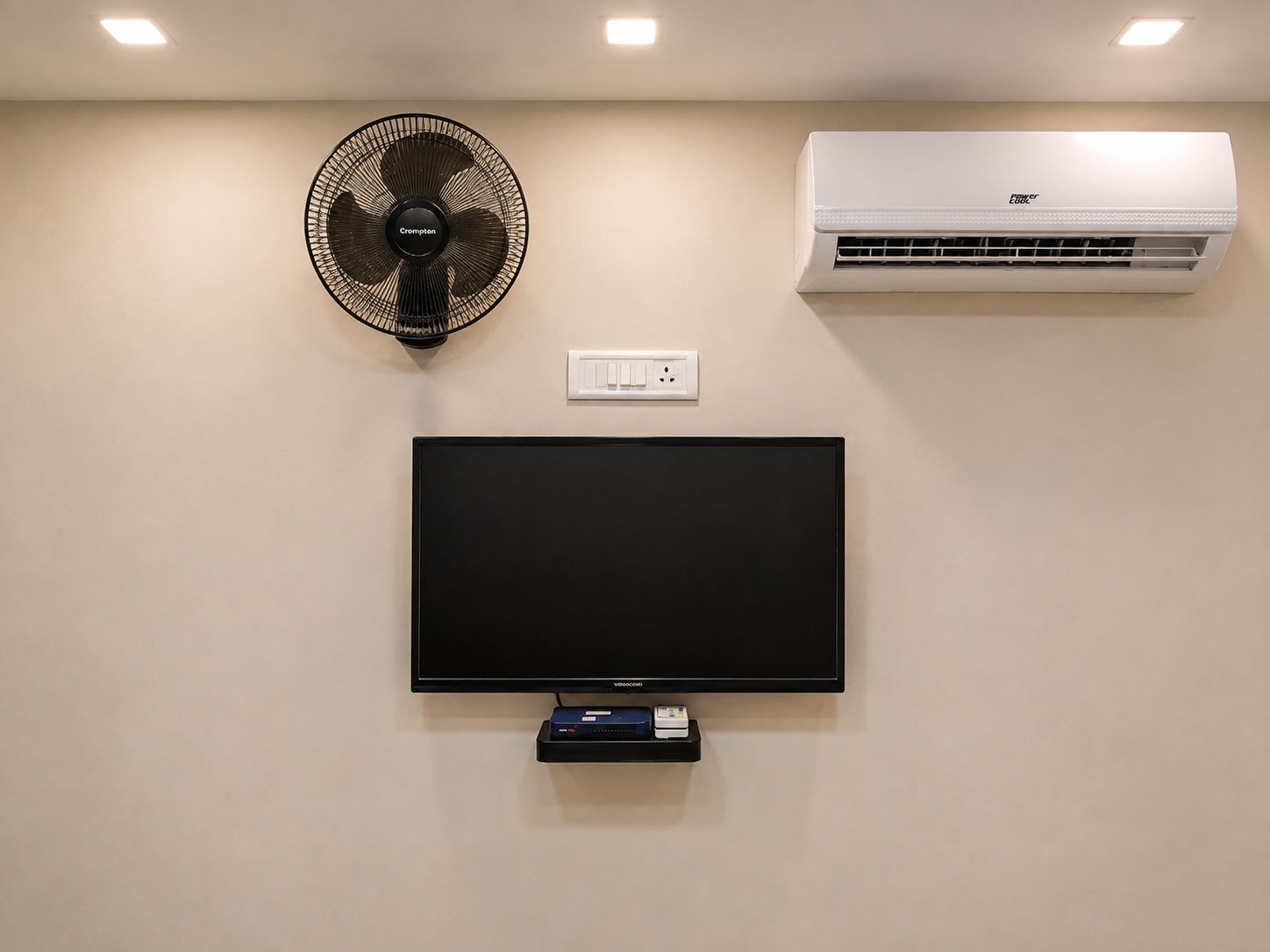 A wall-mounted flat-screen TV, a black standing fan, and a white ceiling-mounted air conditioner.