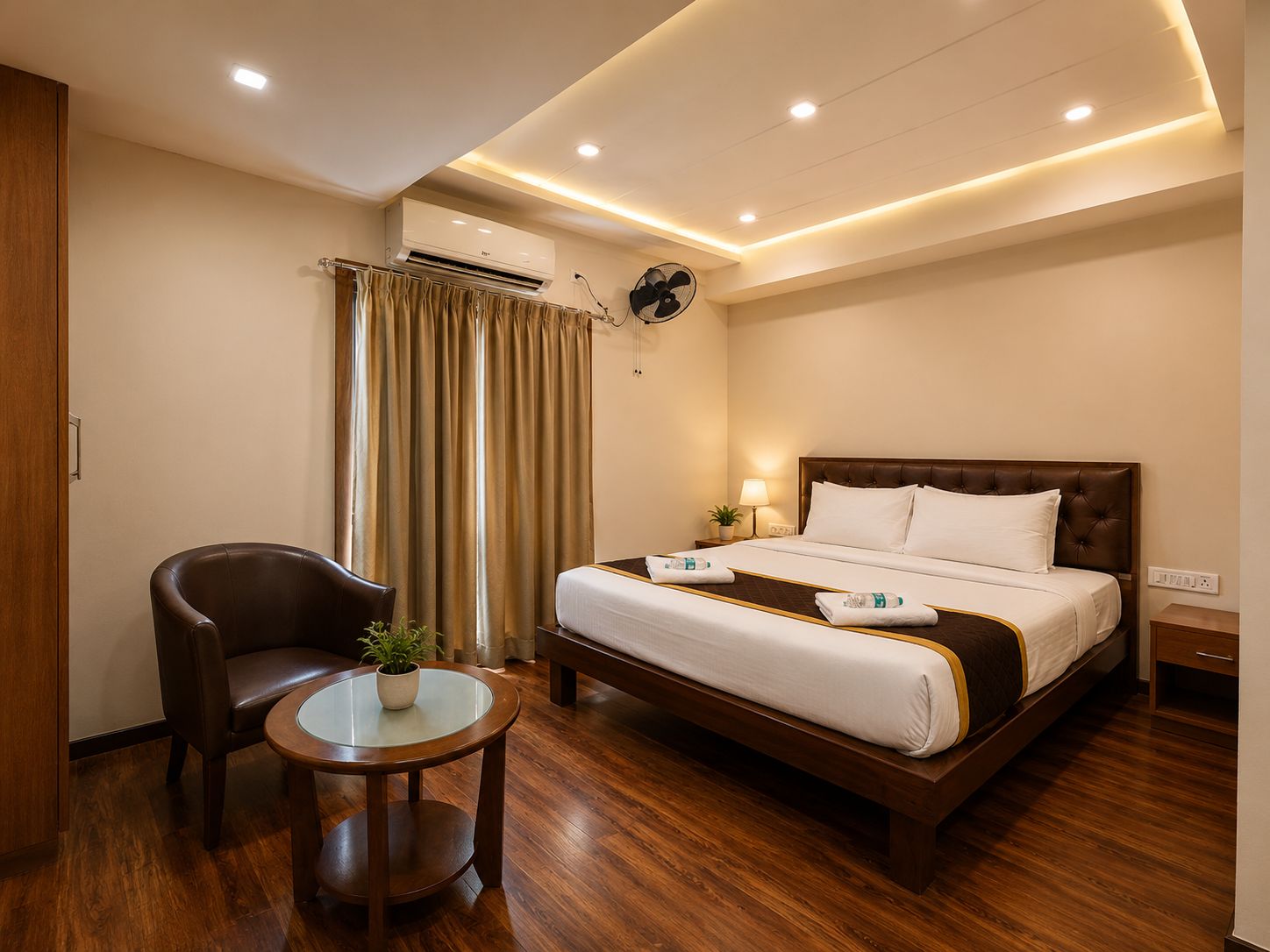 A hotel room with a bed, a chair, a small table with a plant, and a ceiling fan.