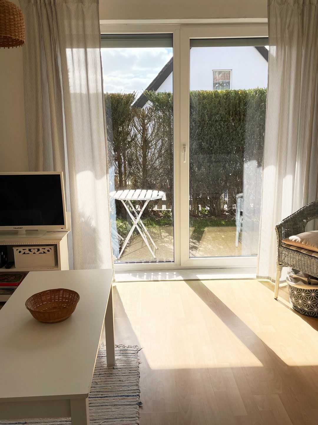 Une pièce avec un téléviseur, une table blanche, une chaise en osier et une porte-fenêtre coulissante donnant sur un jardin.