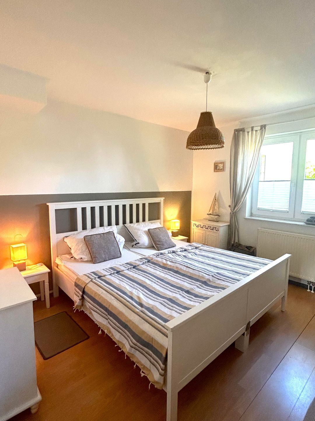 A bedroom with a bed, bedside tables, a lamp, a hanging light fixture, a window with curtains, and a radiator.