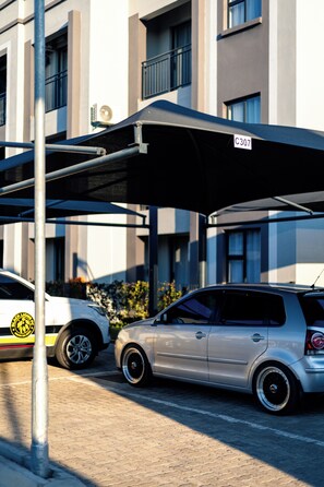 A parking area with a covered carport, a building with multiple windows, and a pole with a sign.