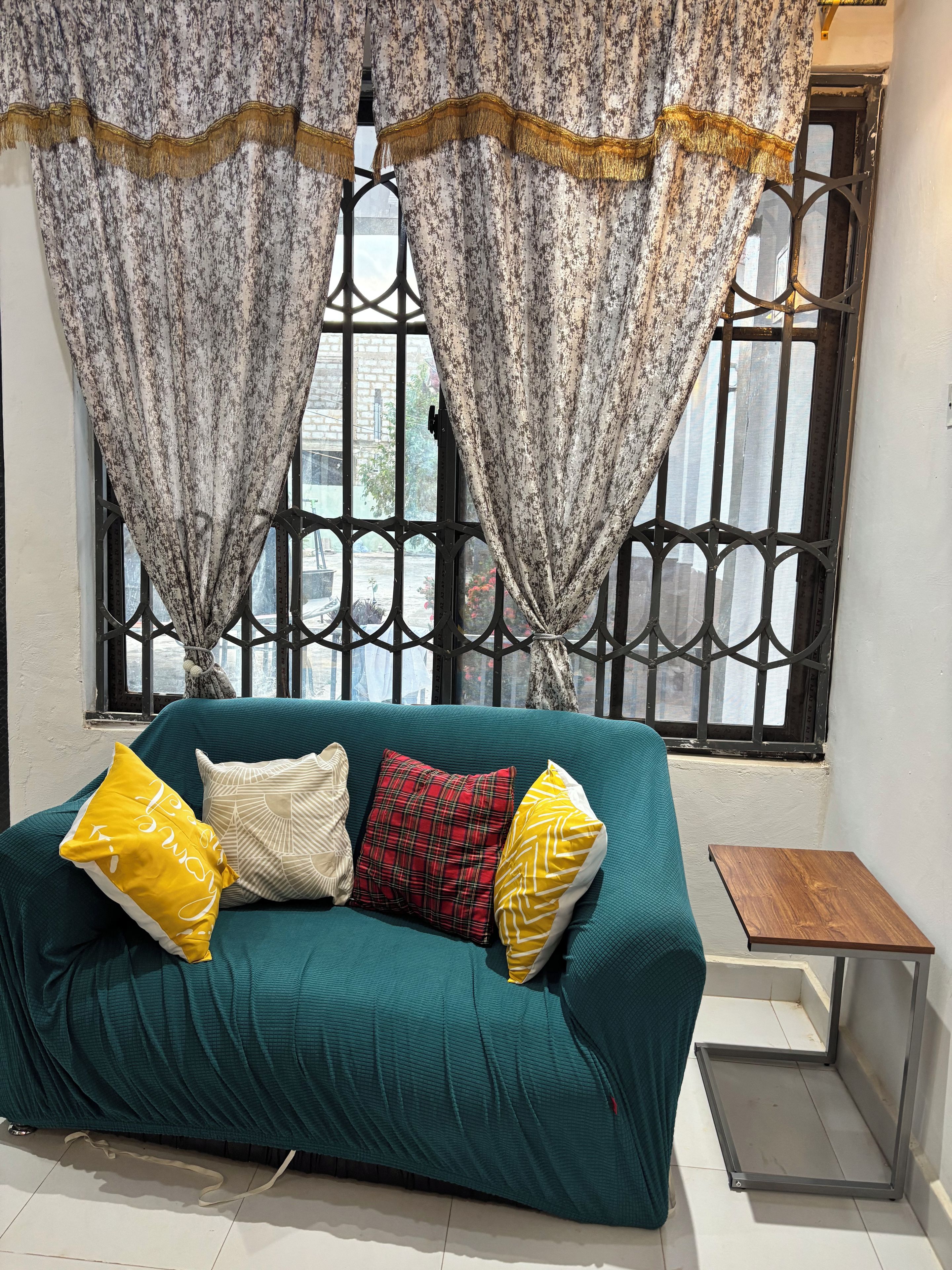 A teal sofa with patterned cushions, a small wooden side table, and a window with decorative curtains.