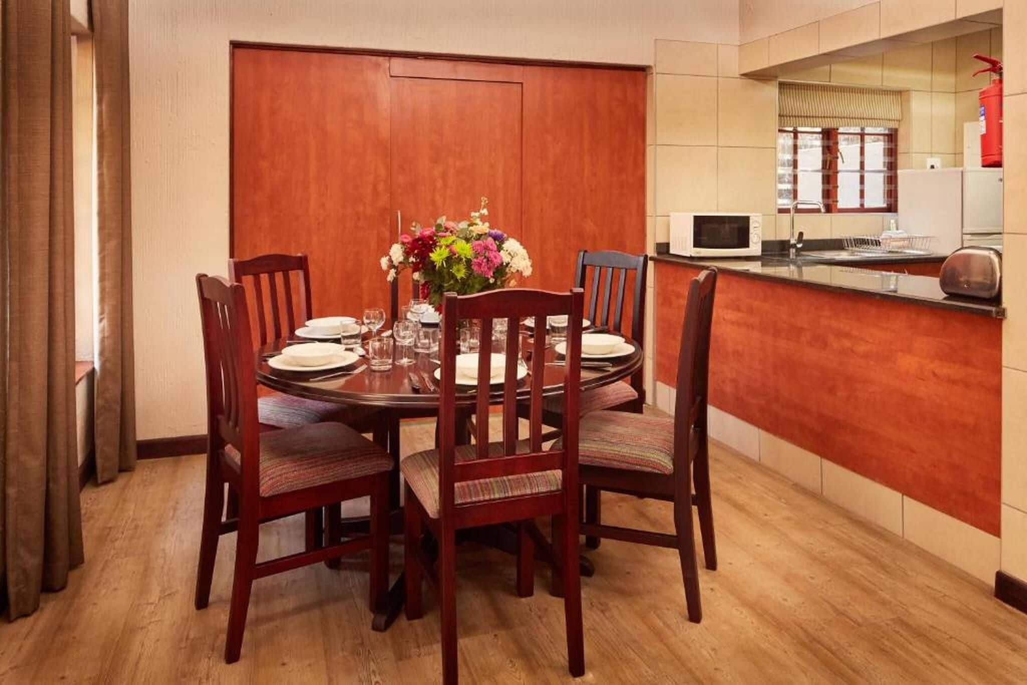 Un comedor con una mesa redonda, cuatro sillas, un jarrón de flores y una zona de cocina con microondas y tostadora.