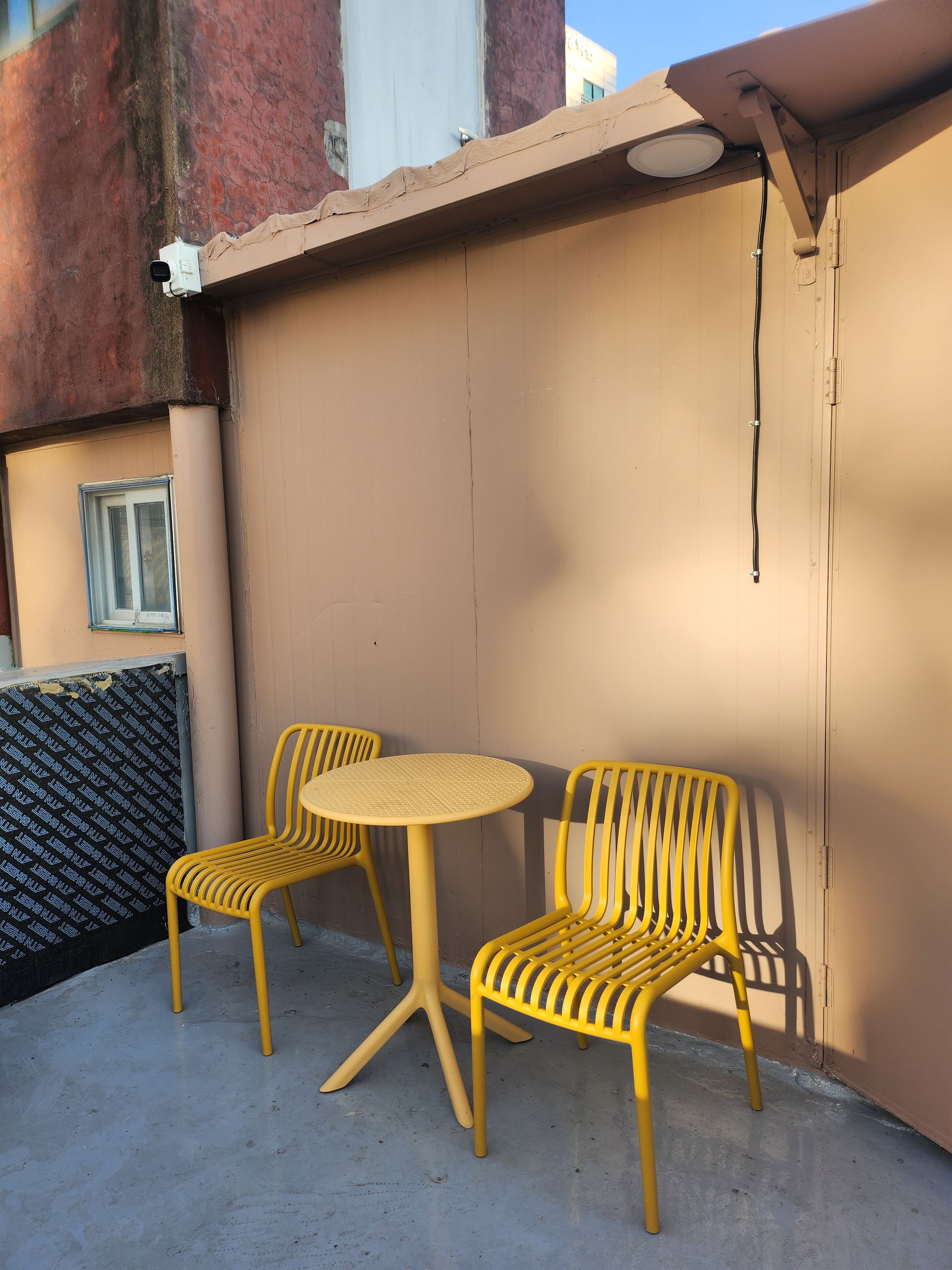 A small outdoor seating area with two yellow chairs and a round table, adjacent to a building with a textured wall and a window.