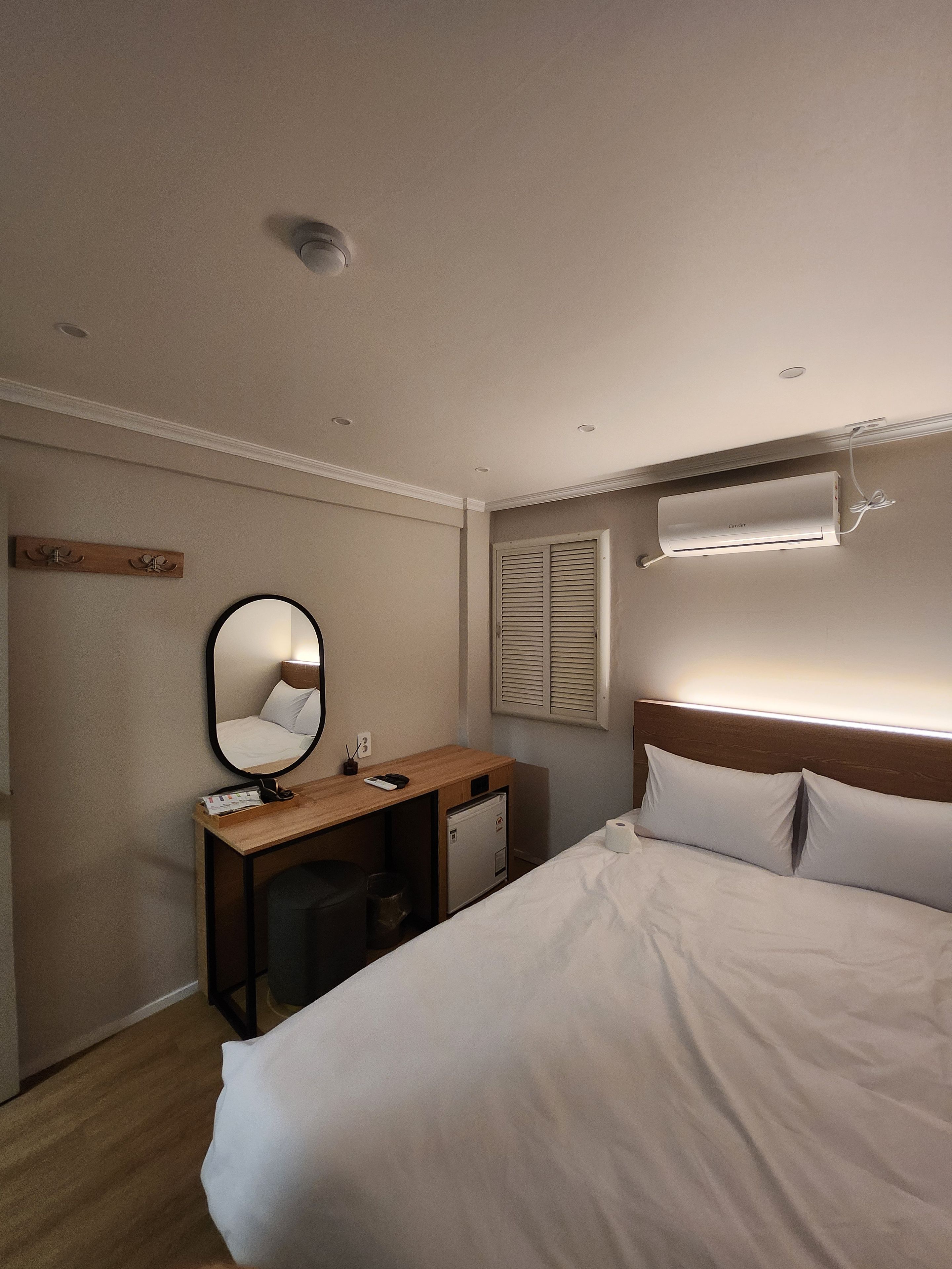 A hotel room with a bed, a desk, a mirror, a wall-mounted air conditioner, and a window with blinds.