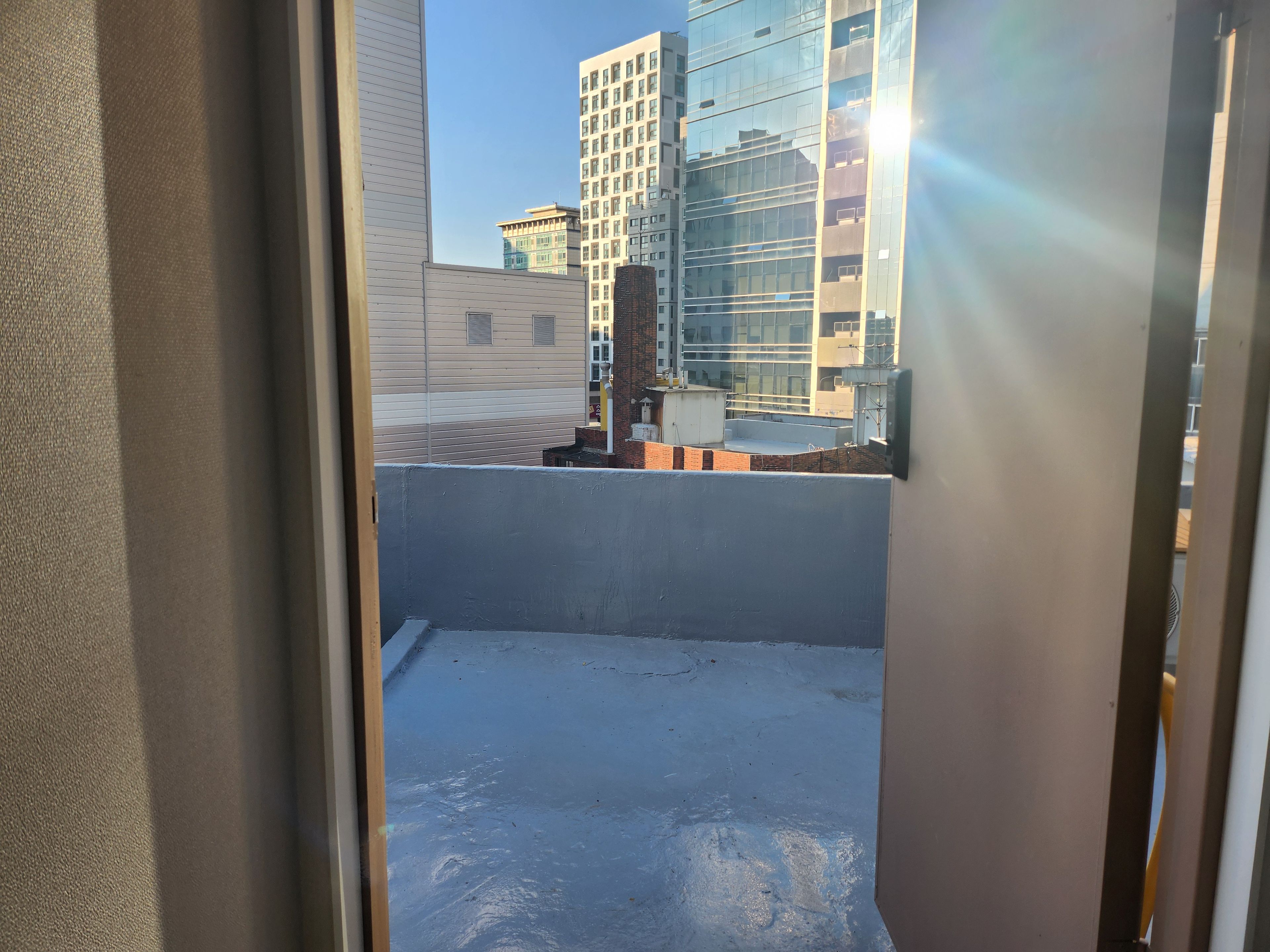 A view through a window with a metal frame, looking out onto a rooftop with a blue surface, buildings in the background, and a clear sky.