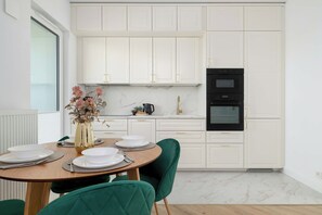 A modern kitchen with white cabinetry, a black oven, and a dining area with a round table and green chairs. - Warsaw | Jagiellońska 79 | Stylish Studio | Balcony (Warszawa)