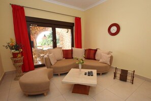 Ein Wohnzimmer mit einem beigefarbenen Sofa, einem Couchtisch, einem roten Vorhang, einer Wanduhr und Blick durch eine Glastür.