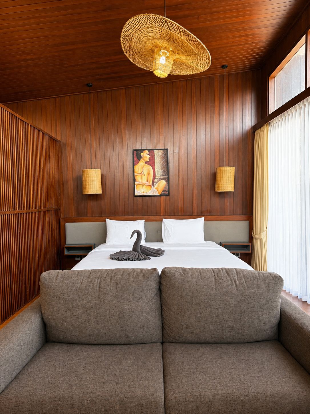 A hotel room with a wooden paneled wall, a bed with white linens, two wall-mounted lamps, and a large woven ceiling light.