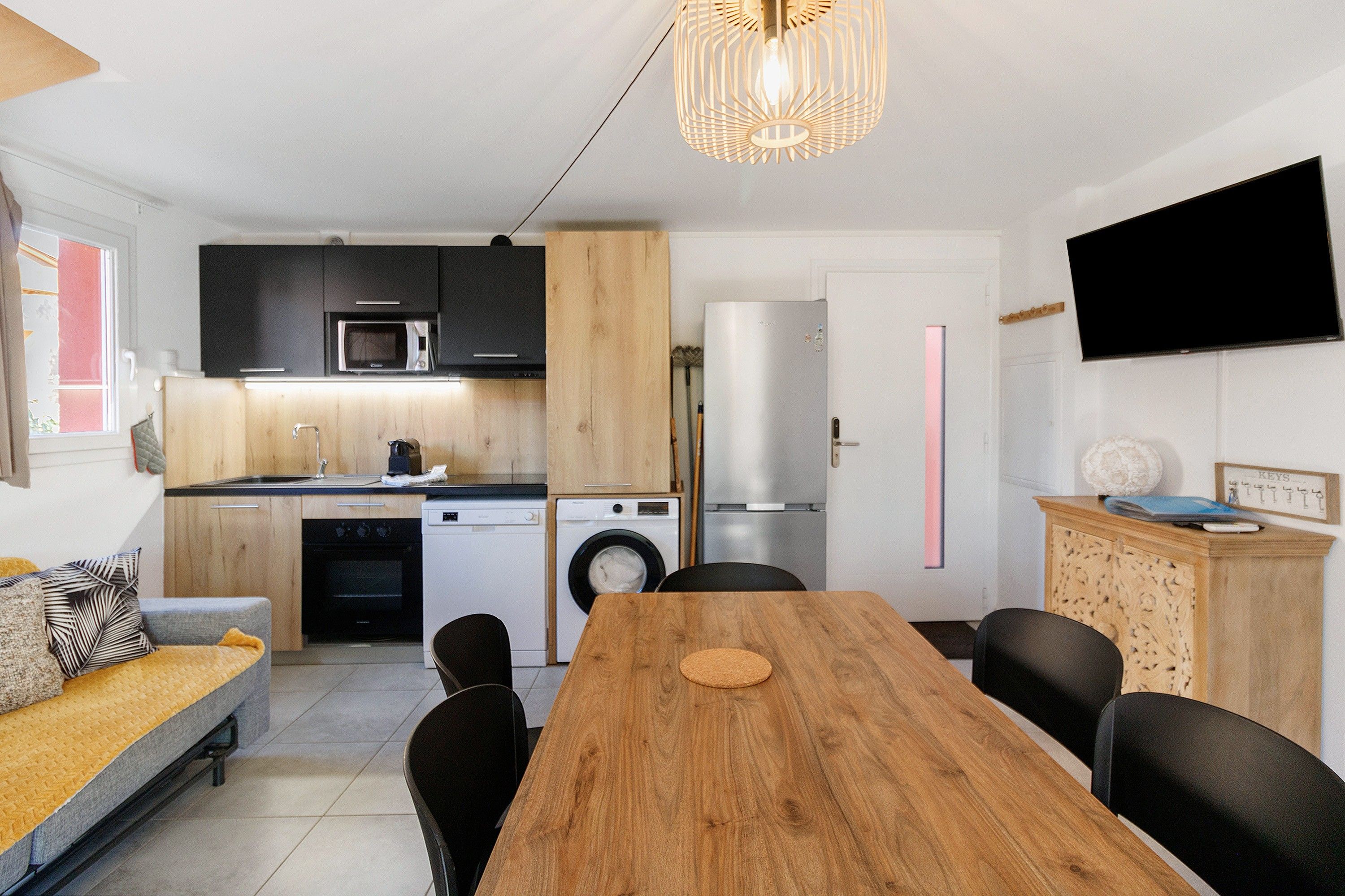 Eine kompakte Küche mit modernen Geräten, ein Essbereich mit Holztisch und ein an der Wand befestigter Flachbildfernseher.