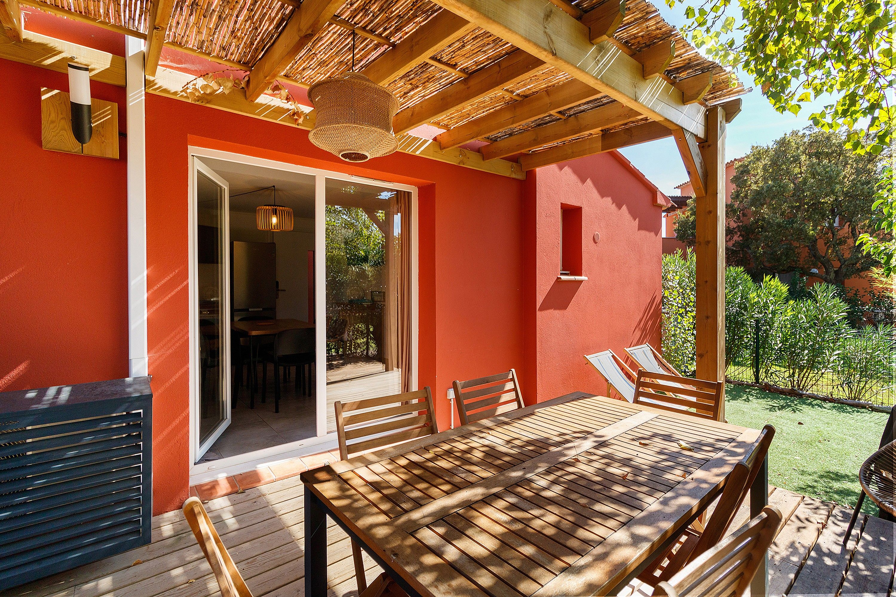 An outdoor patio with a wooden table and chairs, a pergola with bamboo roofing, and a red wall.