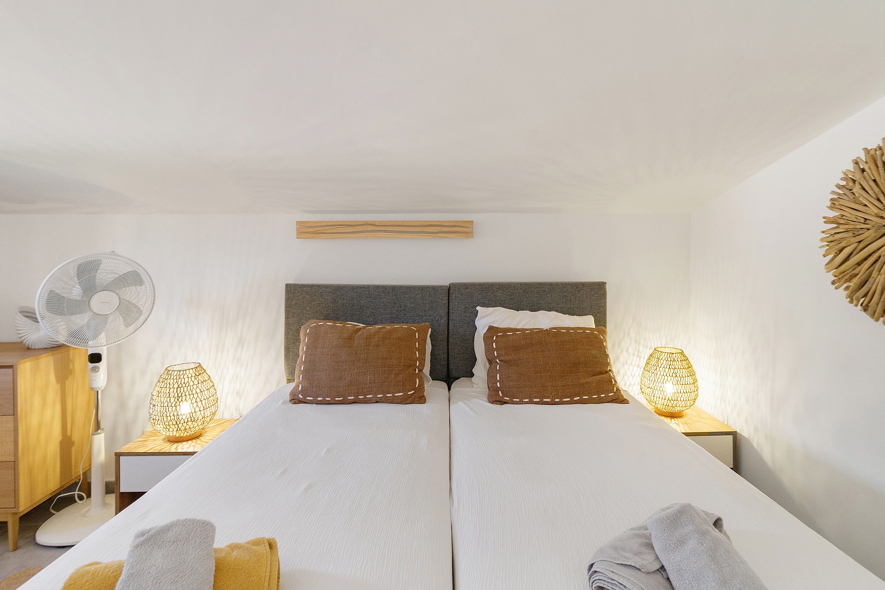 Un letto matrimoniale con due cuscini, un comodino con lampada, una ventola a muro e un elemento decorativo a parete.