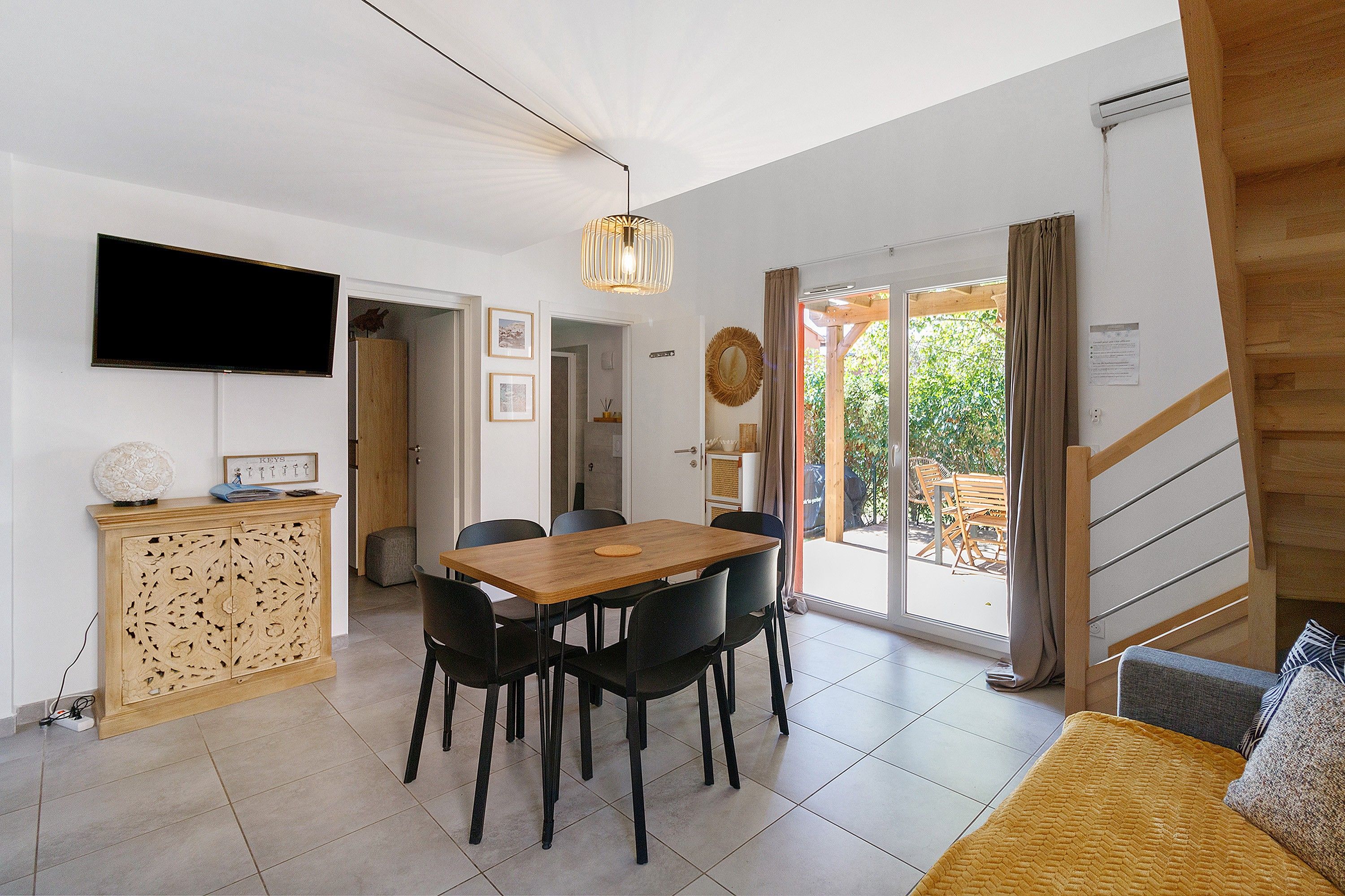 A dining area with a wooden table and chairs, a television mounted on the wall, and a staircase leading to an outdoor area.