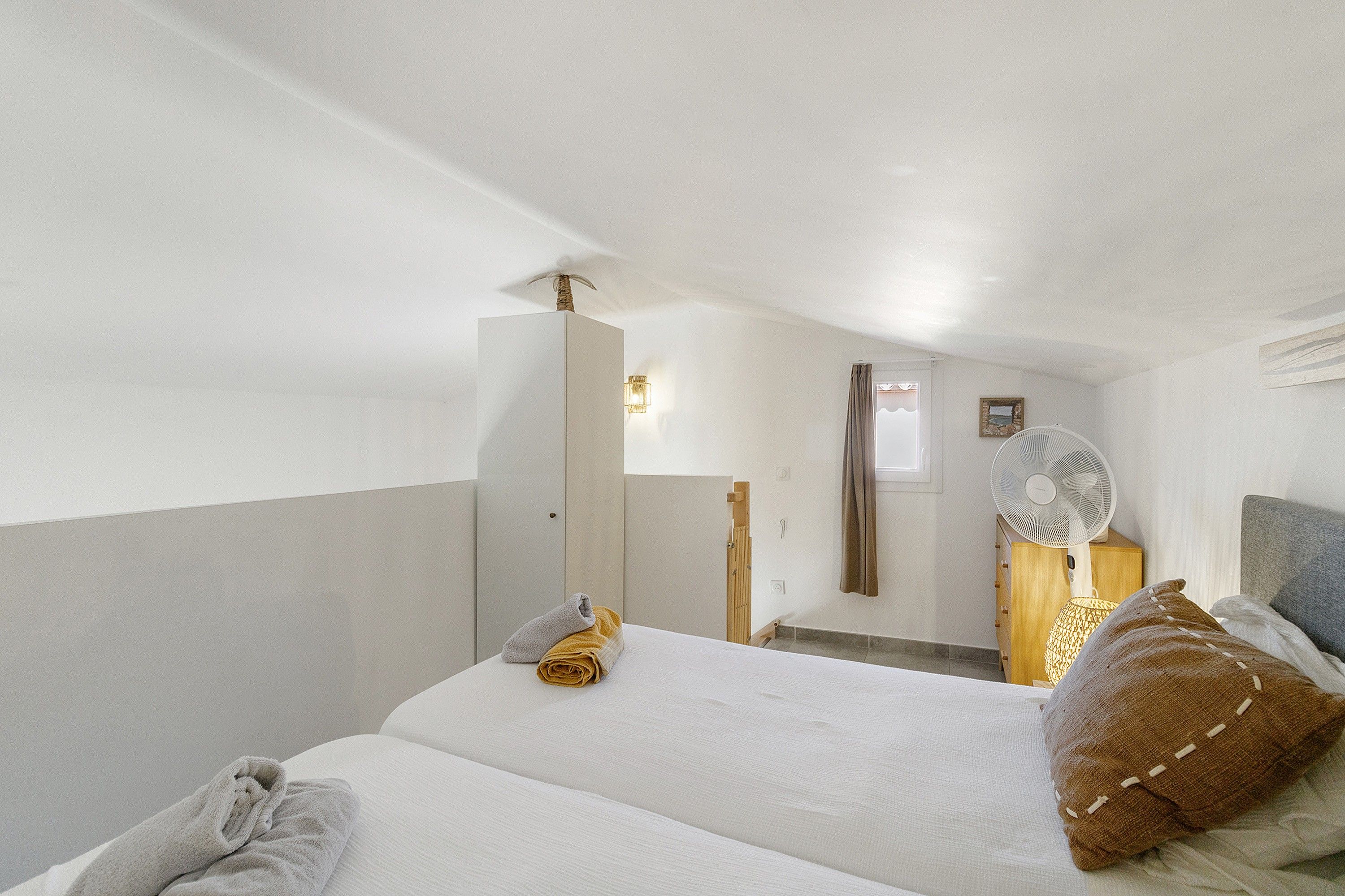 A hotel room with a bed, a bedside table, a mirror, a fan, and a window.