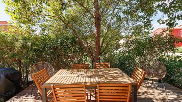 An outdoor wooden table and chairs set up under a tree, surrounded by greenery.
