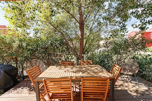 Une table et des chaises en bois, installées à l’extérieur sous un arbre, entourées de verdure.