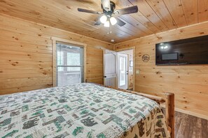 Un dormitorio con techo de madera, una cama con cubrecama estampado, un ventilador de techo y un televisor montado en la pared.