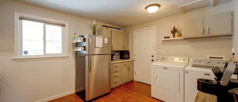 A kitchen with a stainless steel refrigerator, a built-in oven, a washing machine, and a dryer. There is a window with blinds, a door, and a shelf with plants.