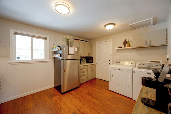 A kitchen with a stainless steel refrigerator, a built-in oven, a washing machine, and a dryer. There is a window with blinds, a door, and a shelf with plants.