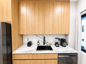 A modern kitchen with wooden cabinets, a marble countertop, a black sink, a coffee maker, and a toaster. - Cozy Chic 2BR – New Kitchen, Complimentary Parking (Miami)