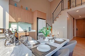 A modern dining area with a marble table set for four, a kitchen in the background, and a staircase leading to an upper level. - Cassia Residence Rama 9 Bangkok (Krung Thep Maha Nakhon)