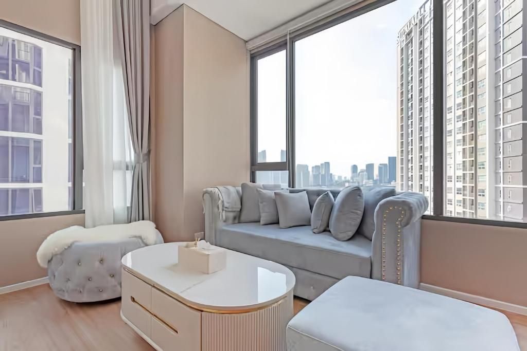 A modern living room with a sofa, ottoman, and a coffee table, large windows with a city view, and a high-rise building in the background.