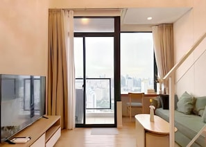 A modern living room with a flat-screen TV, a sofa, a dining table, and a view of the city through large windows. - Cassia Residence Rama 9 Bangkok (Krung Thep Maha Nakhon)