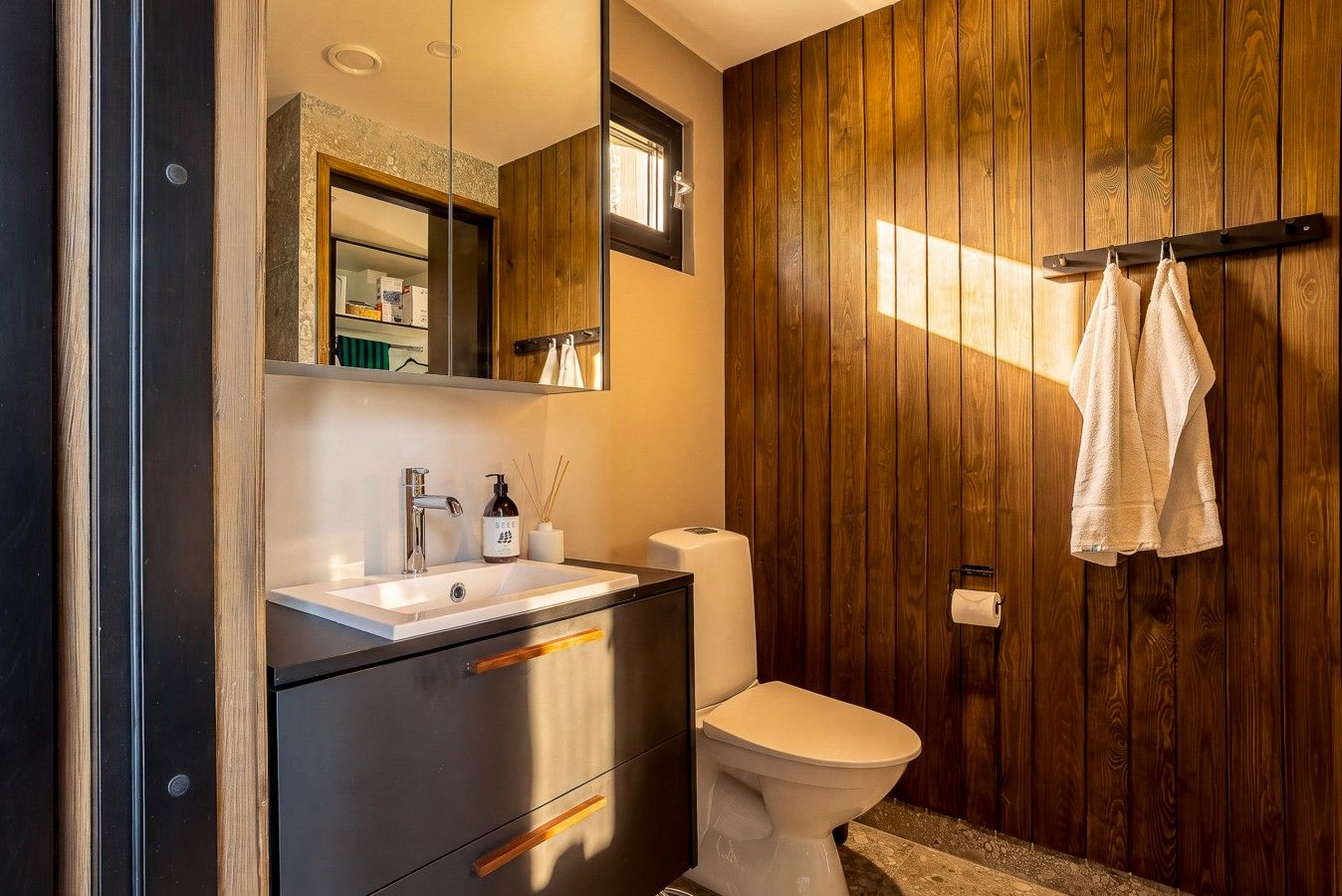 Une salle de bain moderne avec un mur recouvert de panneaux en bois, des toilettes blanches, un lavabo avec un miroir et un porte-serviettes.